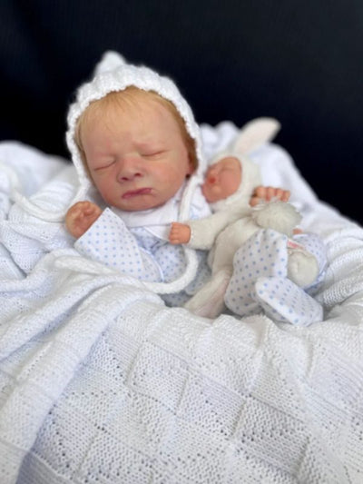 Side view of Reborn Doll Milan lying down with its favorite toy, showcasing a highly visible nose with delicate, sensitive skin details, ultra-realistic facial features, and lifelike Genesis paint.